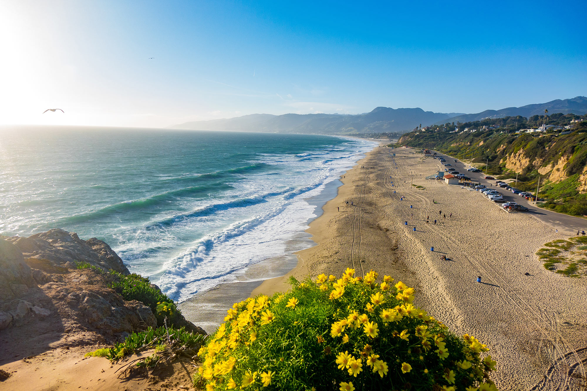 A stunning aerial view of Zuma Beach in Malibu, California, captured golden hour with sand and white-capped waves rolling onto shore.